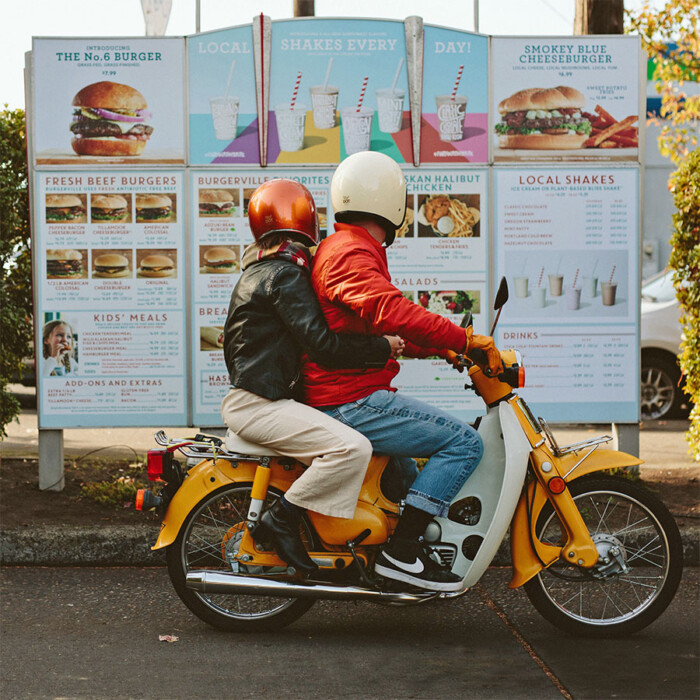 Two people on a yellow vintage motorcycle are reading the outdoor menu for the Burgerville drive-through.