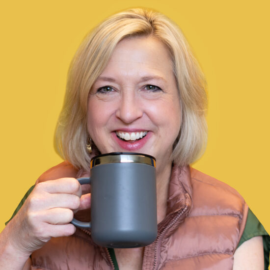 A smiling woman with blond hair in a down vest holding a camping coffee mug.