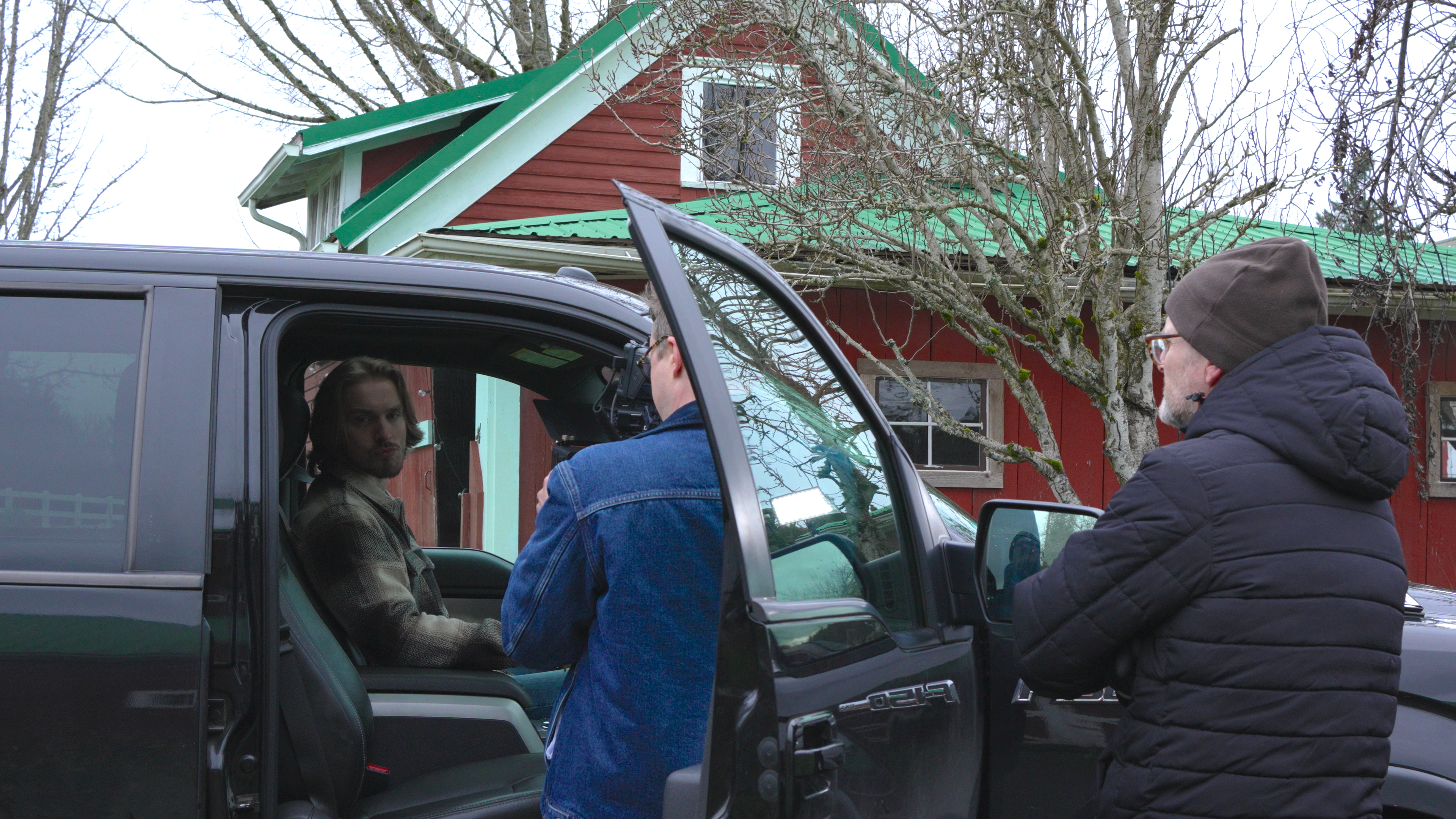 A videographer and a director standing outside a pickup with an actor inside behind the wheel.