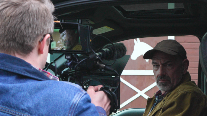 A man holds a video camera and films another man sitting inside a pickup truck. 