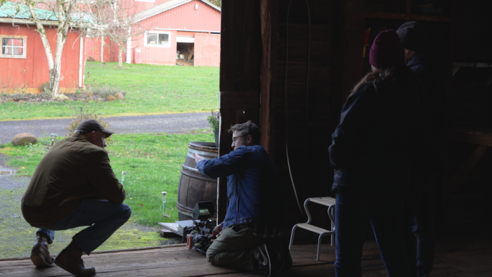 A cameraman talks to an actor inside a barn, looking out onto a green yard and red farmhouse. 
