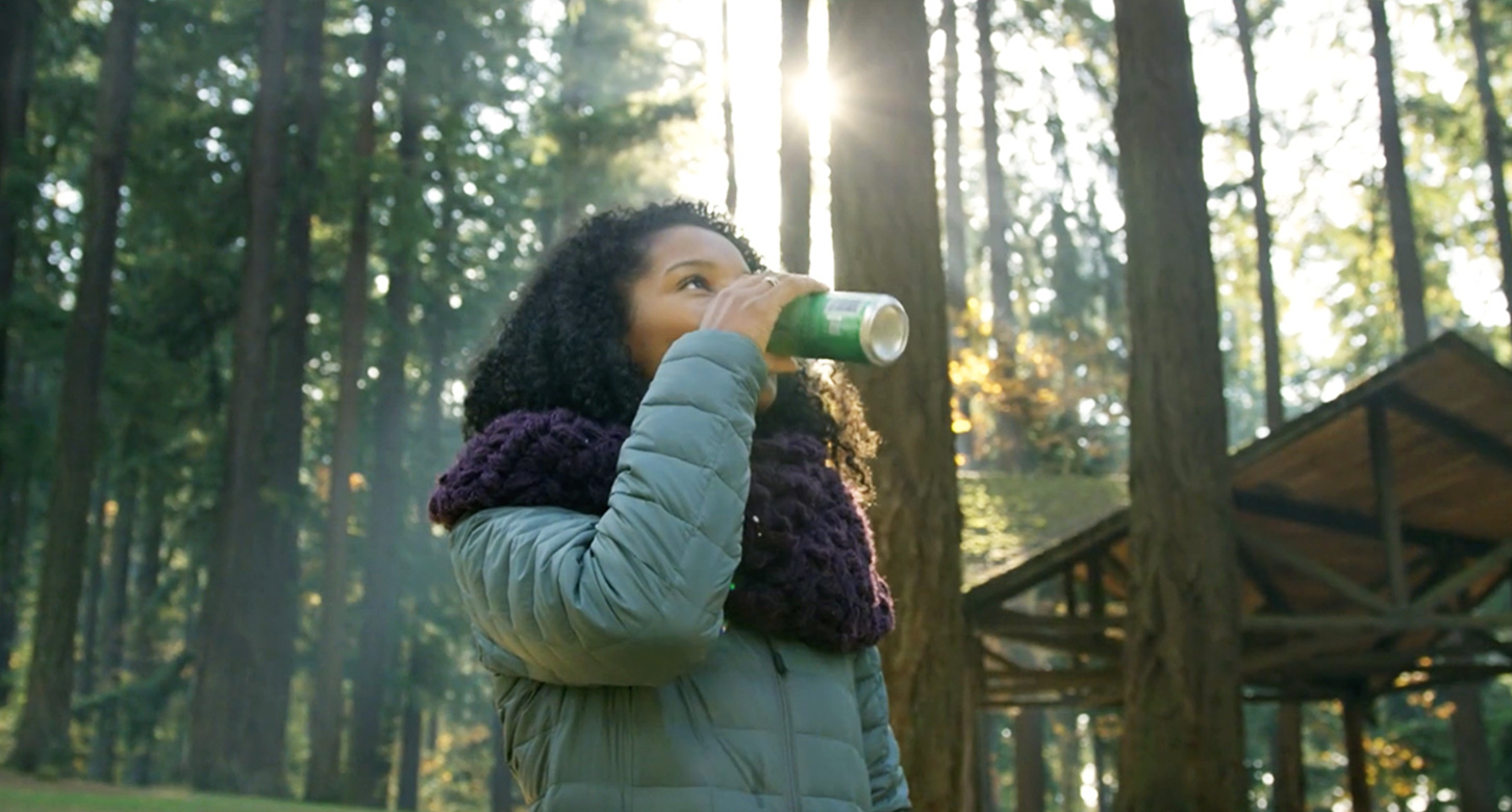 A screenshot from Oregon Beverage Recycling Cooperative's Green Bags video of a woman in a forest drinking a canned beverage.