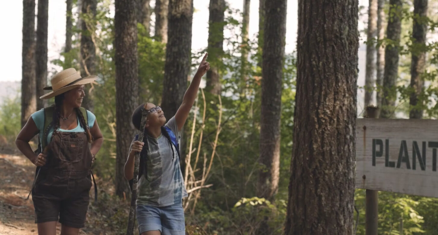 A smiling woman and young girl hike through an Oregon forest while the girl points to something she sees in the trees.