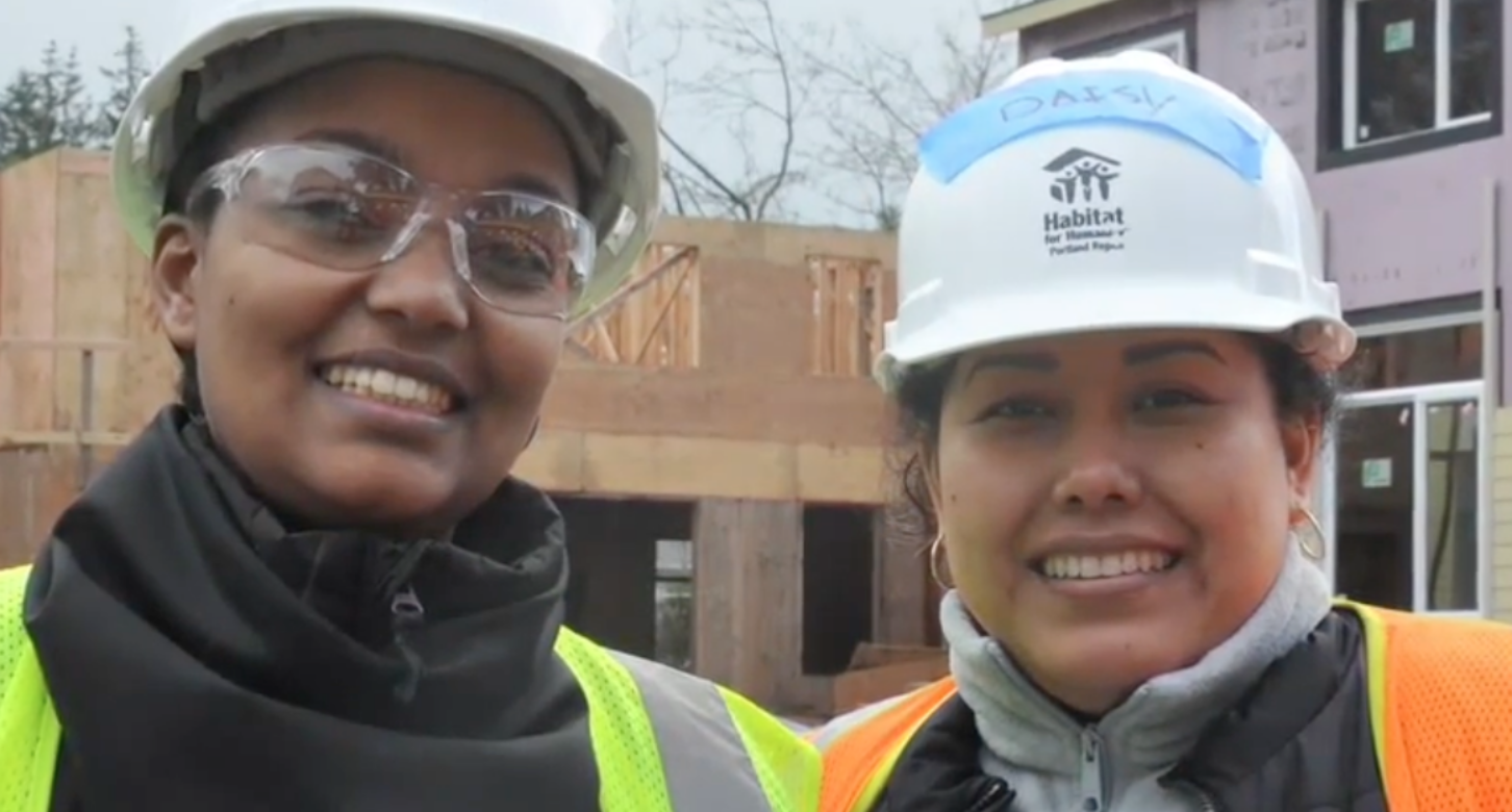 A video still from a news segment about Habitat for Humanity, showing two volunteers outside a home under construction.