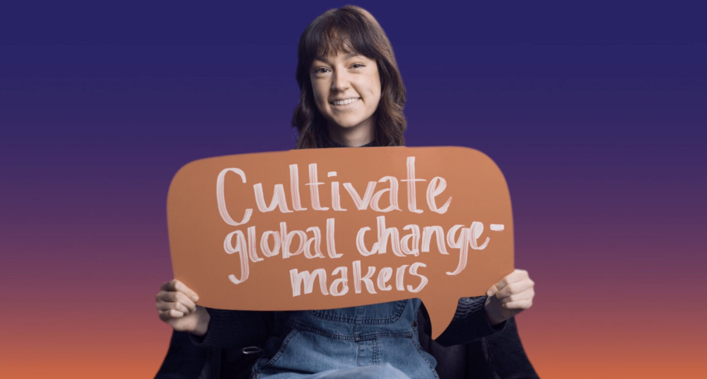 A smiling woman holds a sign in the form of a speech bubble with the words "cultivate global change-makers".