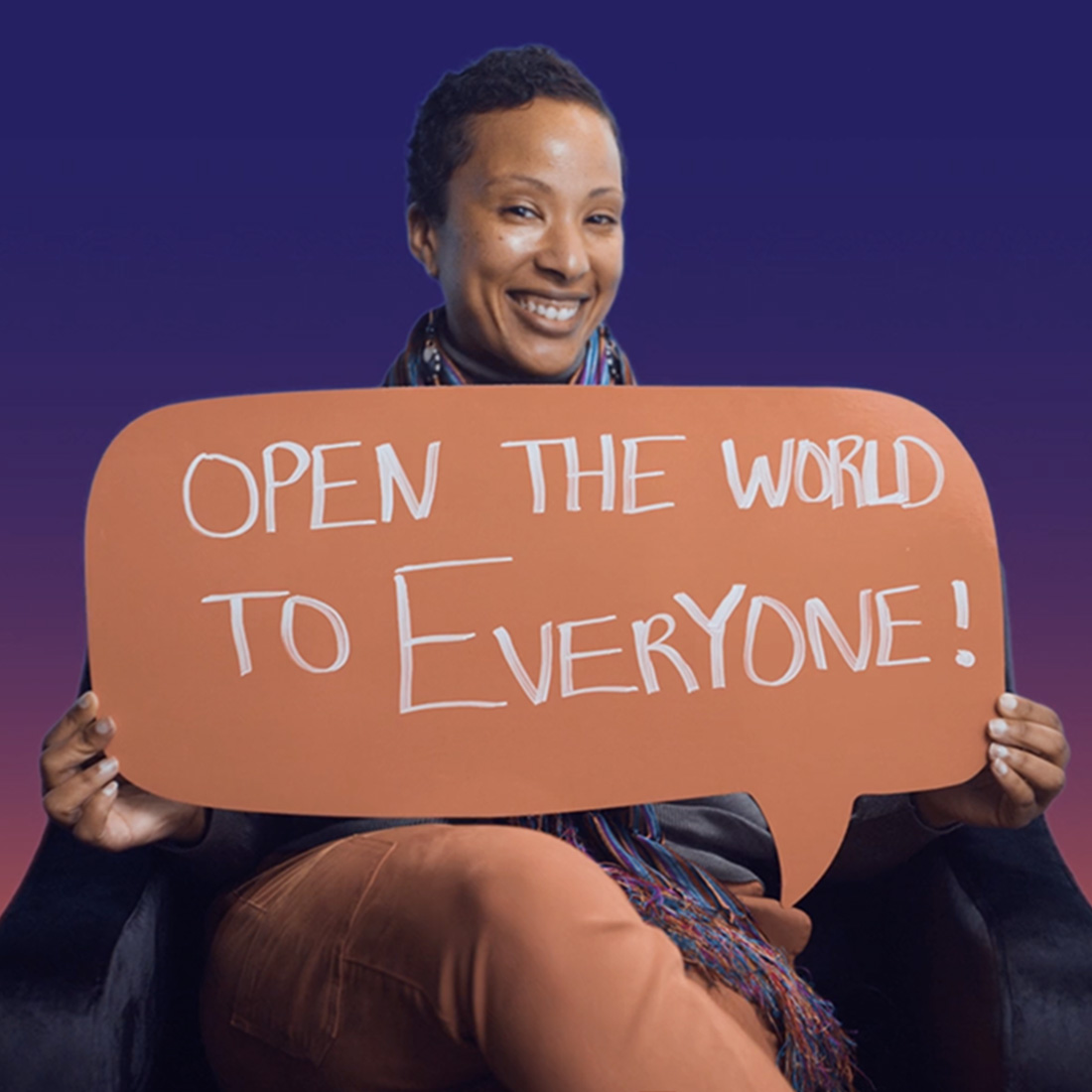 A smiling woman holds a sign in the form of a speech bubble with the words "open the world to everyone!"