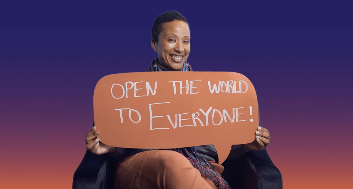 A smiling woman holds a sign in the form of a speech bubble with the words "open the world to everyone!"