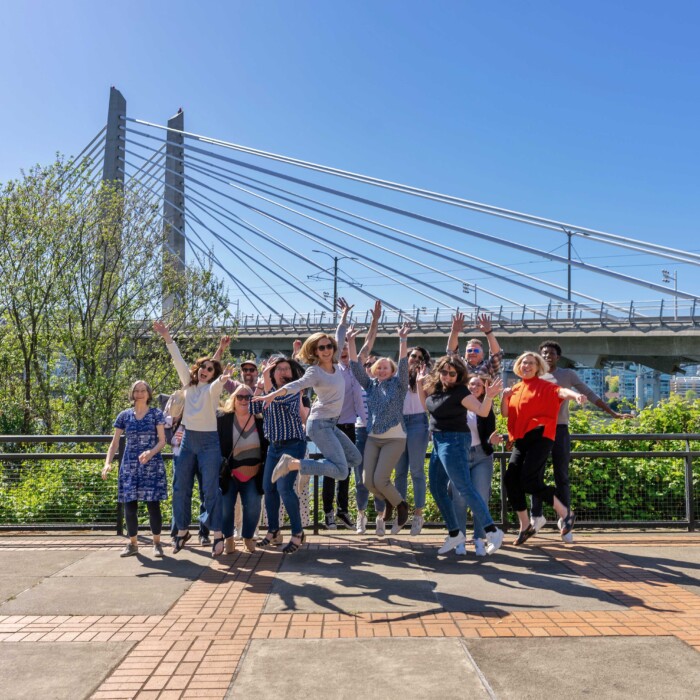 A group of people jump up in the air with their hands raised on a sunny day in Portland.