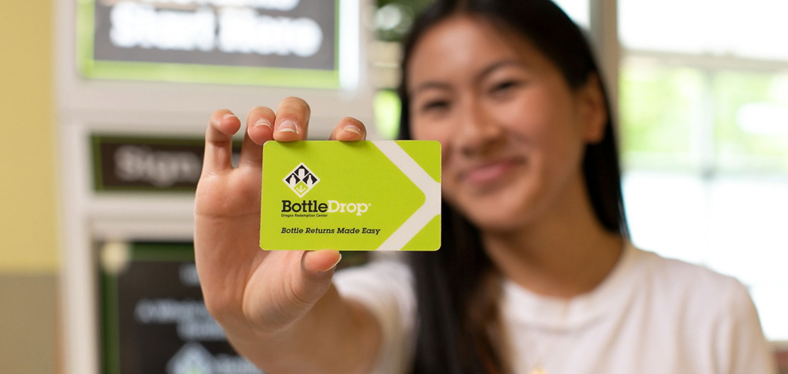 A young woman smiles as she holds out a BottleDrop card for the viewer to see up close.