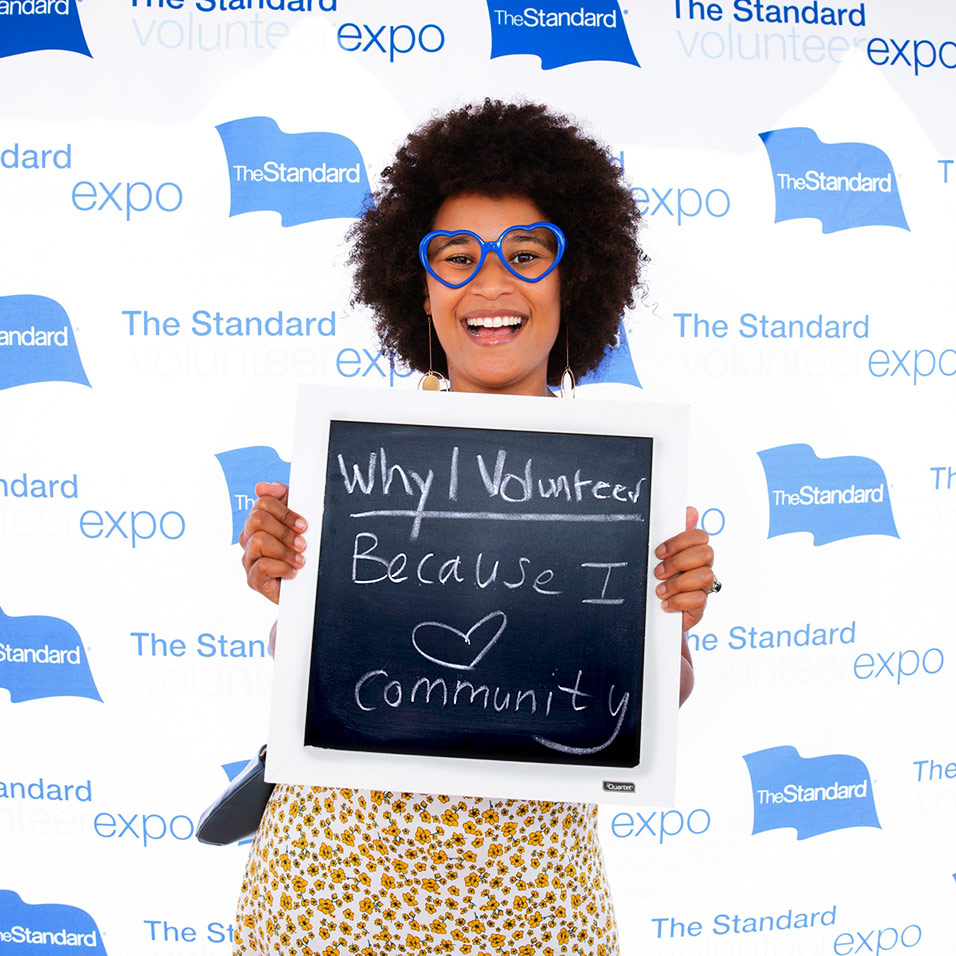 A smiling woman holds a chalkboard sign saying "Why I Volunteer: Because I heart community".