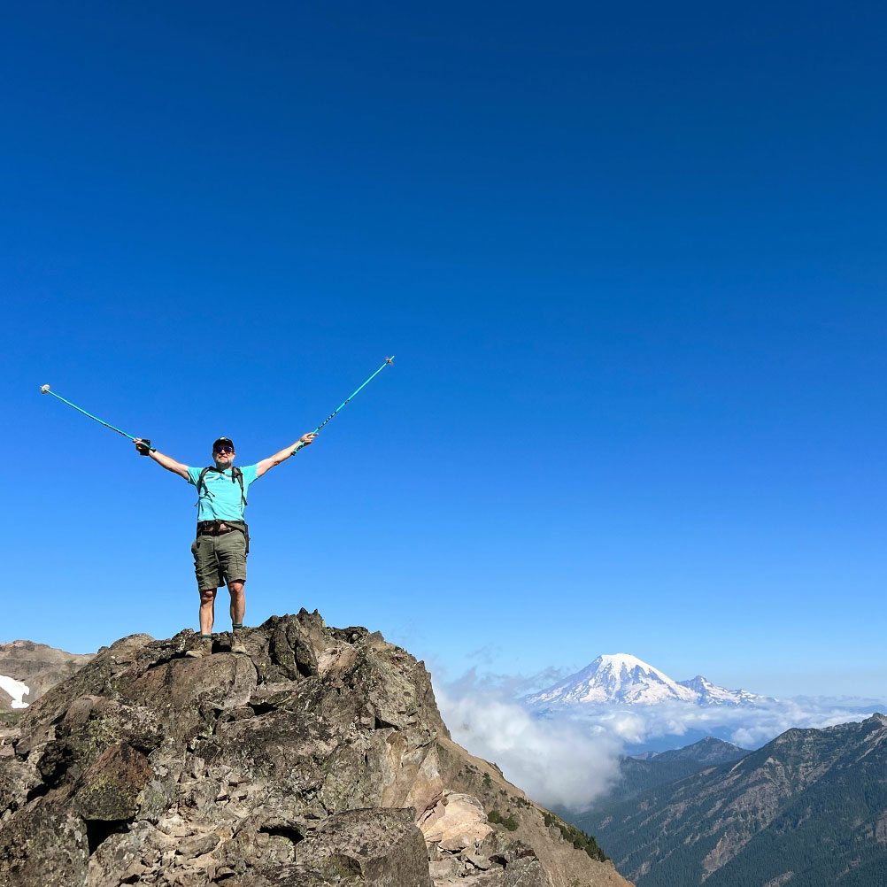 Gard writer Jon Bell celebrates scaling a mountain peak under blue skies in the Pacific Northwest.