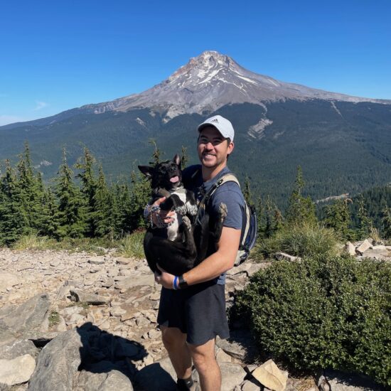 A man holding a dog on a mountainous hiking trail with Oregon's Mt. Hood in the background.