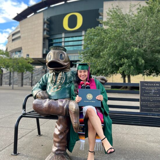 Kery Hernandez holding her diploma and sitting on a bench next to an Oregon Duck statue in front of the UO football stadium.