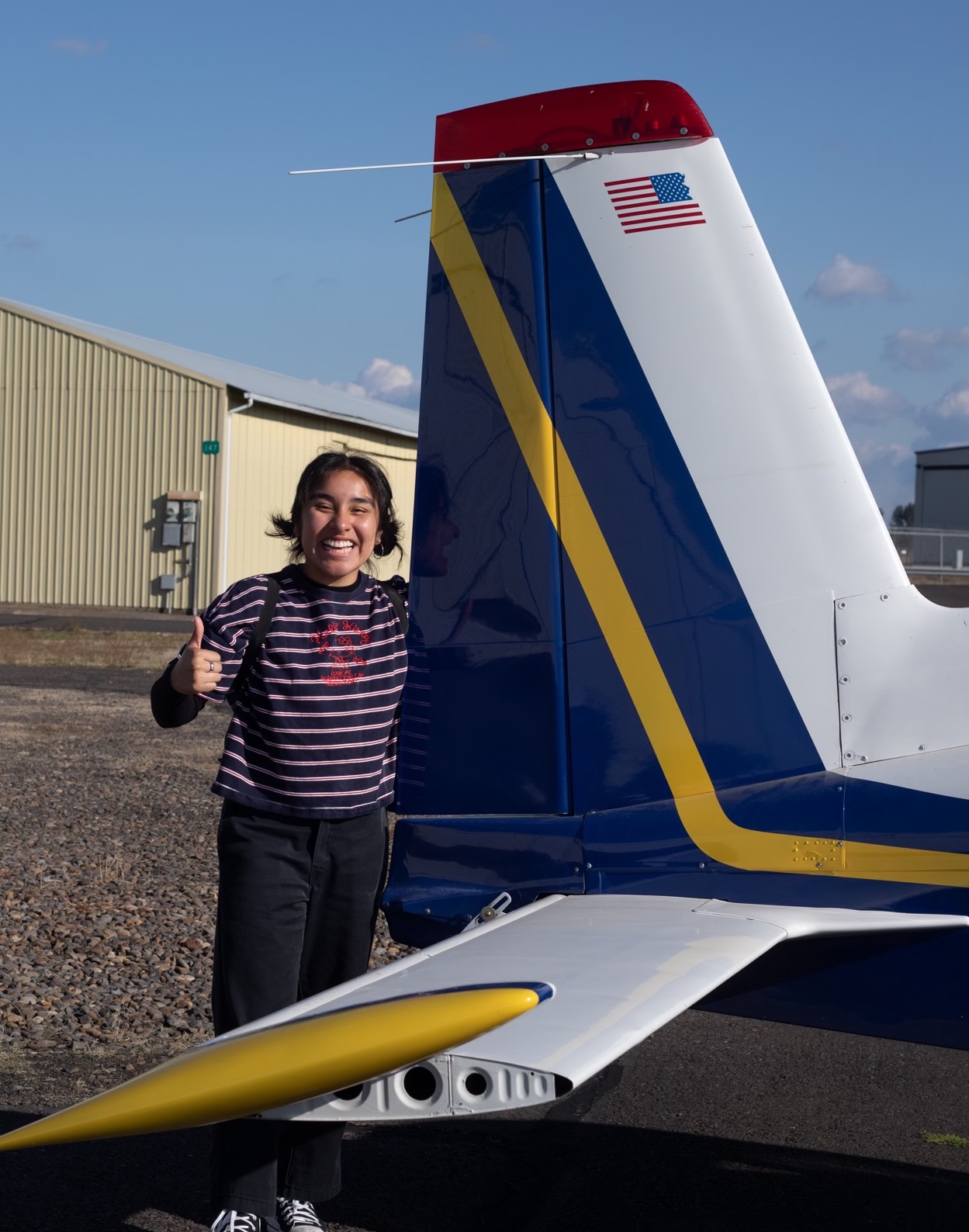 Keyry Hernandez standing next to the tail of a blue, yellow and white airplane on a sunny day.