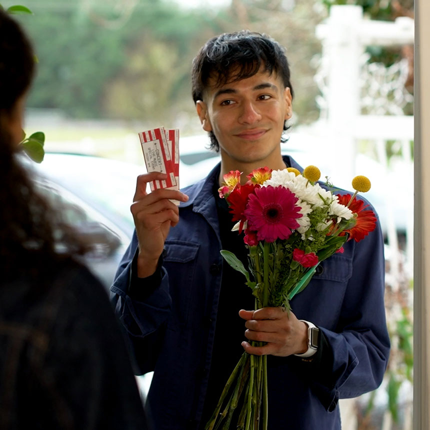 A person at an open door, holding flowers and tickets and looking at a person in the foreground.