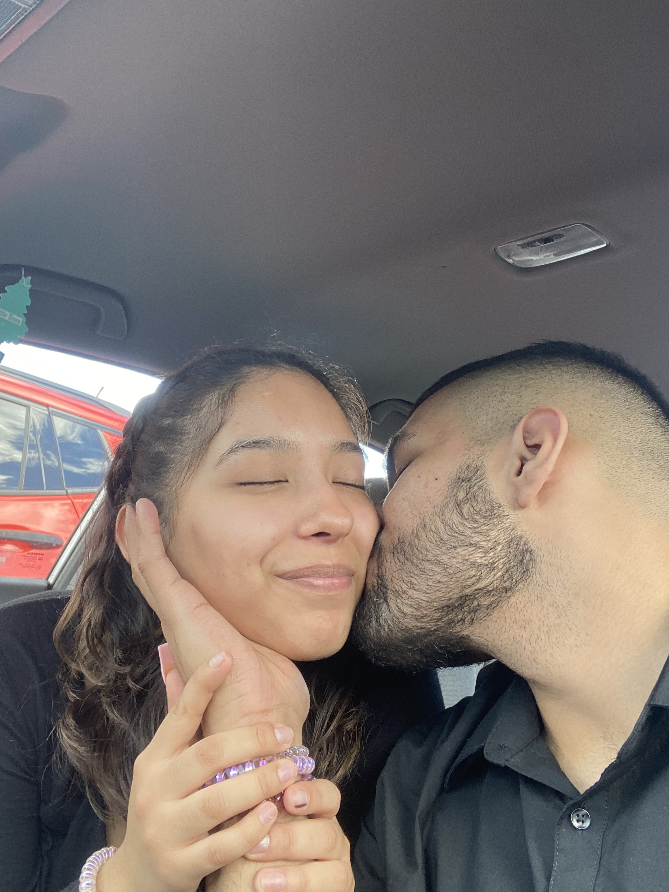 A man kisses his brown-haired girlfriend on the cheek in a car.