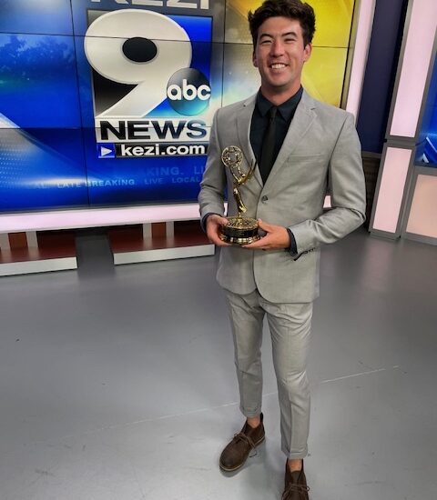 A man in a grey suit holding a gold Emmy Award statue in a TV studio.