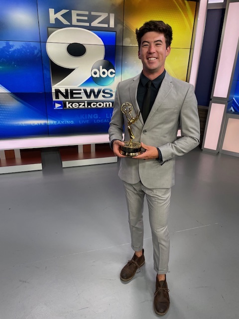 A man in a grey suit holding a gold Emmy Award statue in a TV studio.