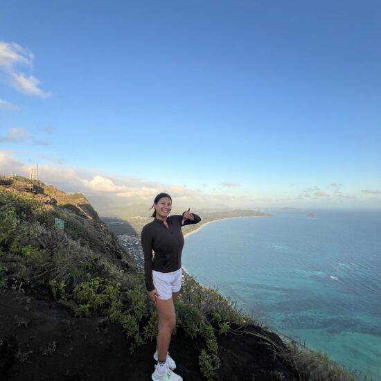 A woman stands among green plants on a hill overlooking the blue waters of Hawaii.