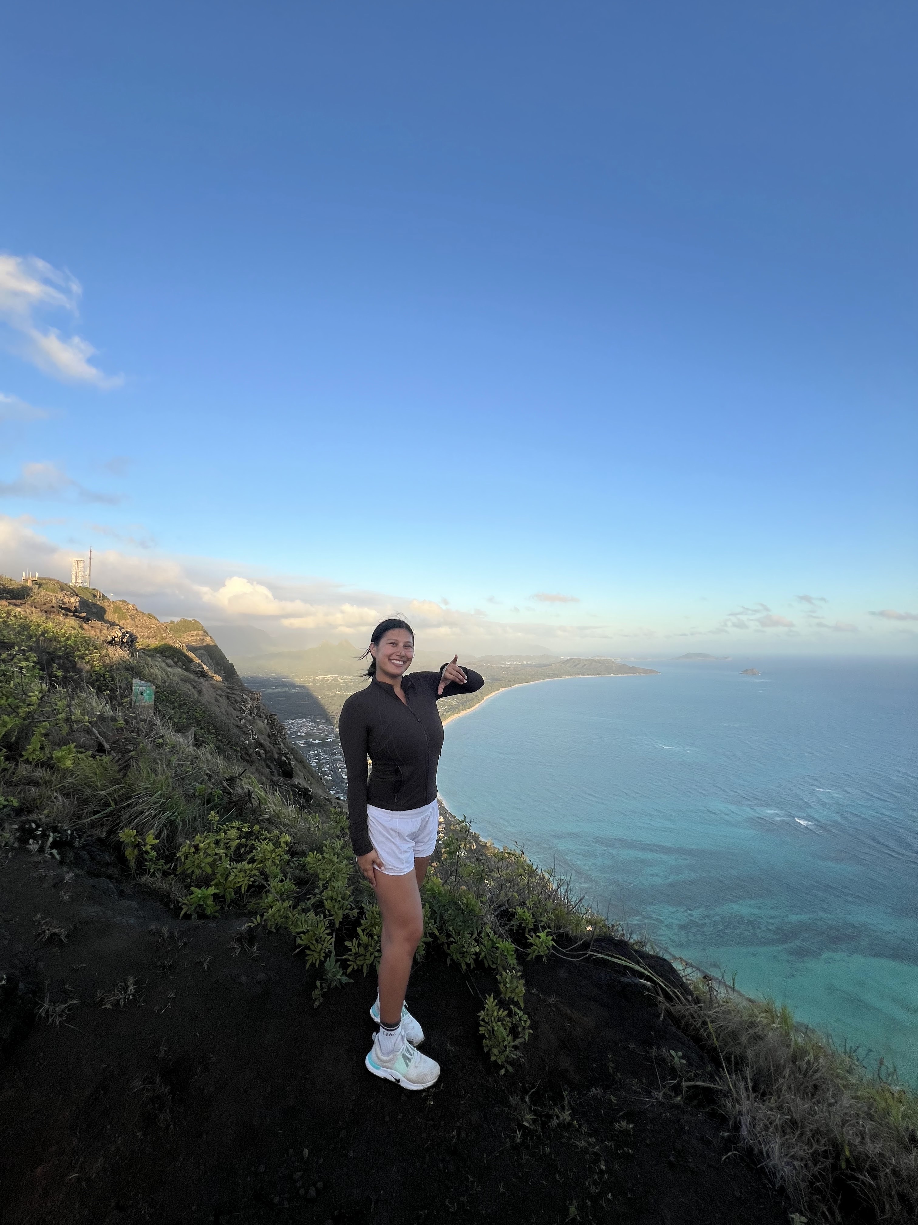 A woman stands among green plants on a hill overlooking the blue waters of Hawaii.