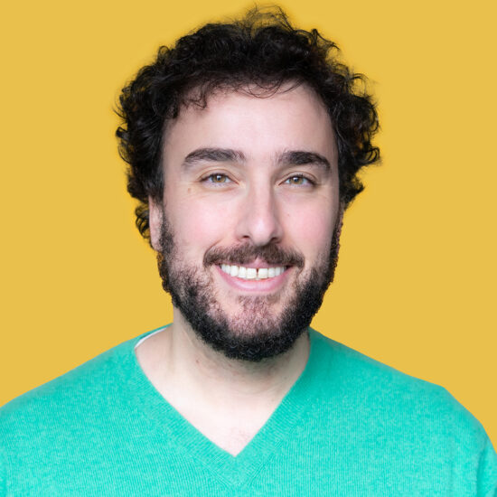 A headshot of a smiling man with dark brown hair and a beard in a green sweater.